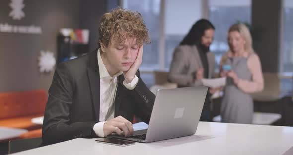 Portrait of Tired Caucasian Young Man Typing on Laptop Keyboard, Looking at Smartphone, and Rubbing