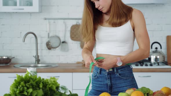 Young girl measures her waist in the kitchen near a table with fruits and vegetables.