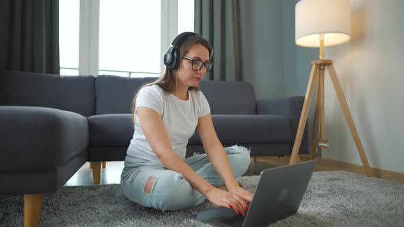 Casually Dressed Woman with Headphones is Sitting on Carpet with Laptop and Working in Cozy Room