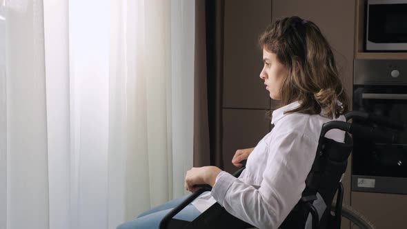 Pensive Disabled Woman Sits in Wheelchair By Window at Home