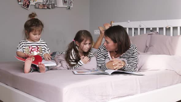 Young Loving Mother and Daughters are Reading a Book and Discussing While Lying on Bed in Bedroom at