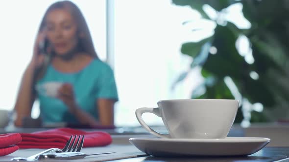 Selective Focus on a Cup on Cafe Table, Woman Talking on the Phone on Background