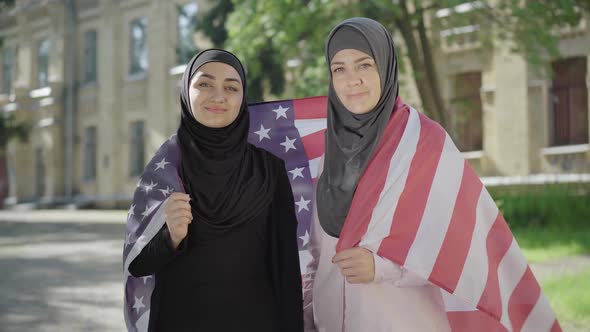 Two Muslim Women Wrapped in American Flag Looking at Camera and Smiling. Portrait of Confident
