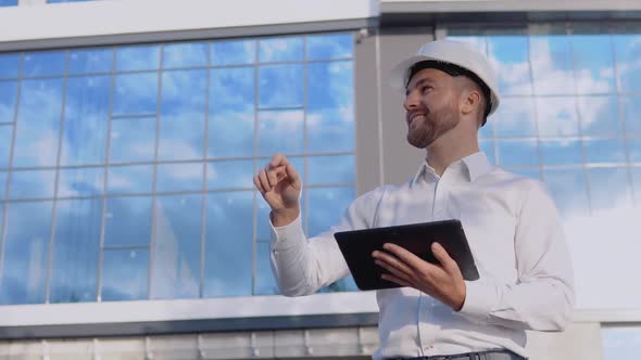 An Engineer Architect in a White Shirt and Helmet on the Background of a Modern Glass Building Works