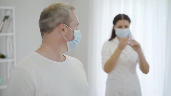 Portrait of Frightened Mid-adult Man in Face Mask Looking Back at Doctor Holding Syringe and Turning