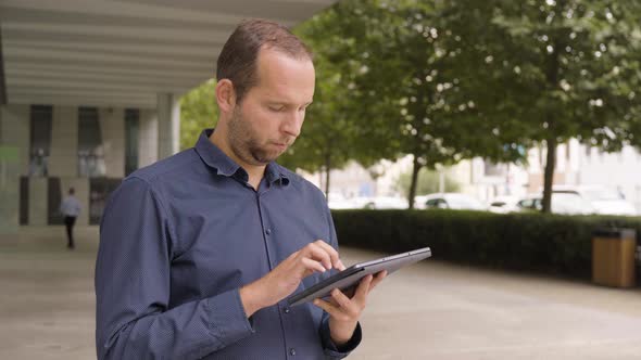 A Caucasian Man Works on a Tablet By an Office Building in an Urban Area