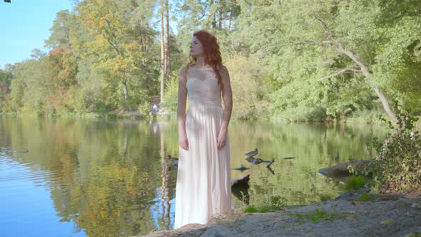Gorgeous Young Caucasian Woman with Red Hair in Long White Dress Stands on the Bank of the Lake