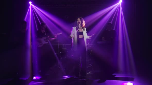 A Stylish Girl Vocalist Sings on Stage in a Vintage Microphone. In the Background Musicians