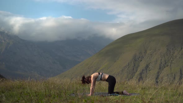 Young Woman in a Tracksuit Is Engaged in Yoga Performing the Pose of a Cat in the Mountains.