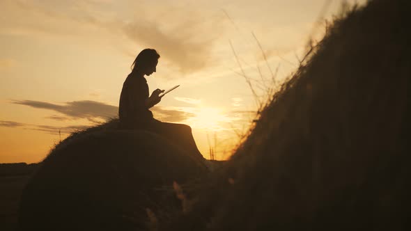 Girl Farmer Uses Tablet in the Field Next To Haystack at Sunset. Smart Farming, Using Modern