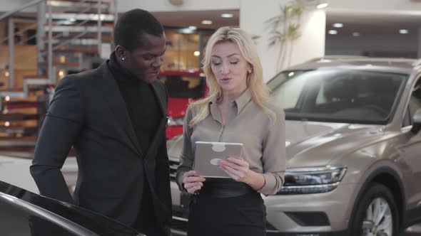 Adult Caucasian Woman with Blond Hair Holding Tablet and Explaining Specifications To African