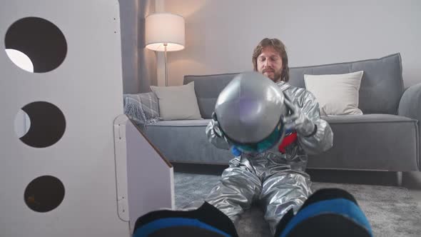 Man in an Astronaut Suit Sits on the Floor in Living Room Next to a Cardboard Model of a Space