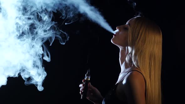 Blonde in a Spacious Room Smokes an Electronic Cigarette. Black Background. Side Viwe