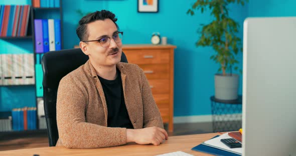 A Man Wearing Glasses with a Mustache Sits in a Company Office in Front of