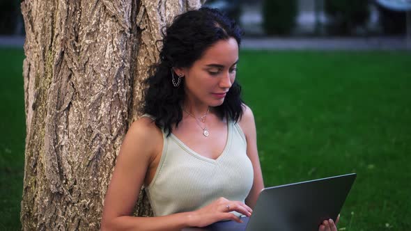 Portrait of a Woman in Dress Sits Below Tree at the Park on Grass Holding Laptop on Her Knees Typing