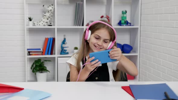 Happy Schoolgirl in Headphones Watching Music Video on Cell Phone in Classroom Study Online