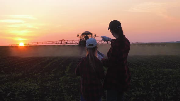 Family Farmers Work in the Field, Look at the Digital Tablet Against the Background of the Tractor