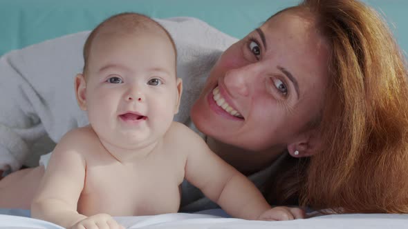 Smiling Beautiful Caucasian Mother Playing with Newborn Baby Boy on the Bed in Bedroom