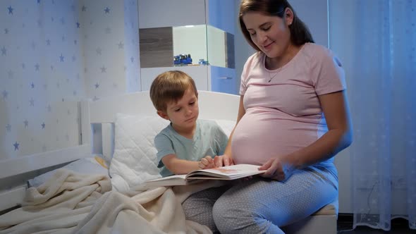 Beautiful Pregnant Mother Redaing Story Book To Her Older Toddler Son Lying in Bed at Night Before