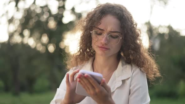 Portrait of Intelligent Concentrated Woman in Eyeglasses Scrolling App on Smartphone in Sunny Park