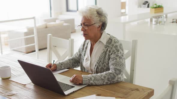 Thoughtful african american senior woman taking notes and using laptop at home