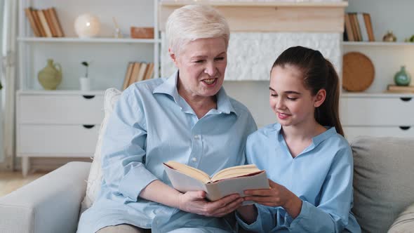 Senior Granny Holding Book and Kid Girl Sitting Together on Couch Indoors