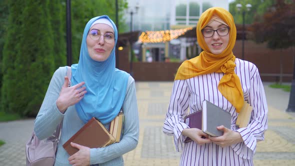 Two Muslim Students in Traditional Scarves with Books in Their Hands Go Through the Park and