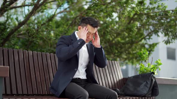 exhausted young asian business man office worker sitting on bench in city park outdoors. 