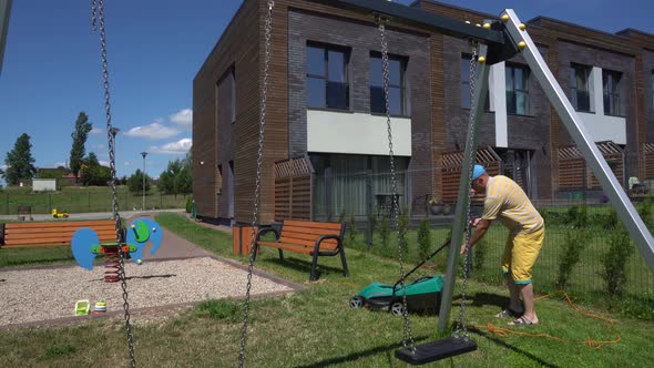 Strong Man in Shorts Cutting Grass Lawn Near Private Children Playground