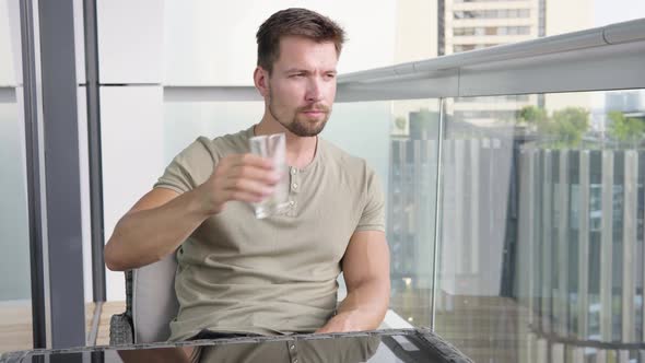 A Young Man Drinks a Glass of Water As He Sits on a Balcony in a Luxurious Apartment and Looks Out