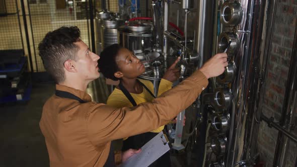 Diverse male and female colleague at gin distillery inspecting equipment and discussing