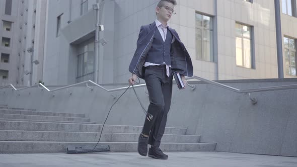 Cute Little Boy Wearing a Business Suit Holds Books in His Hands and Pulls a Computer Keyboard Cord