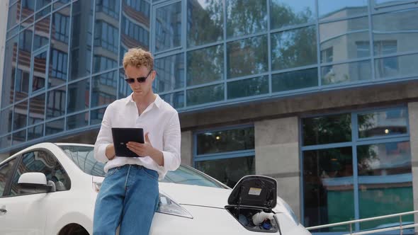 Stylish Modern Young Curly Man Stands Near an Electric Car Charging and Uses His Tablet