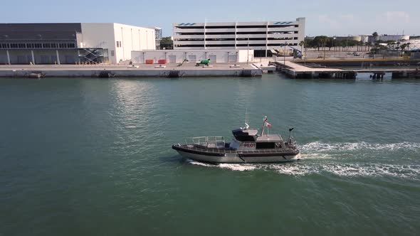 Pilot boat following ship in channel toward open sea with dock, warehouses and other buildings in ba