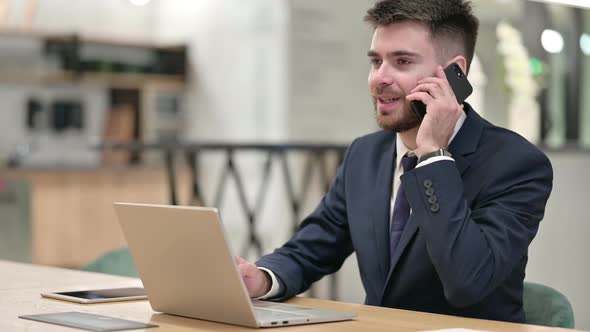 Young Businessman with Laptop Talking on Smartphone in Office