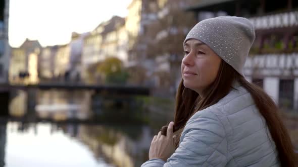 a Portrait in Profile of a Brunette with Long Hair and a Grey Cap Against a Background of Houses, a