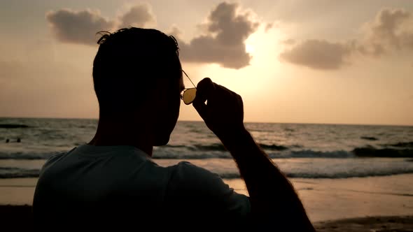 Silhouetted of Man Stands at the Ocean Putting on Sunglasses