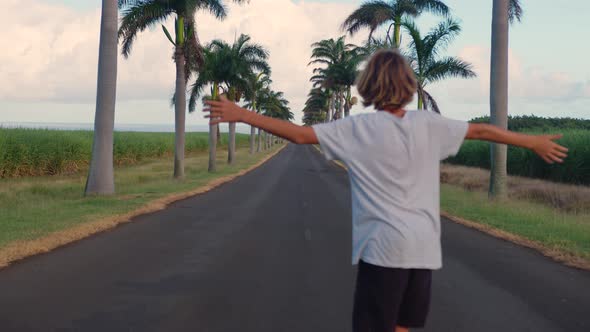 A Teenager with Long Hair Rides a Skateboard Along a Beautiful Road with Palm Trees