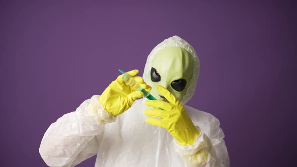 A Man in an Alien Mask in a White Suit Shows a Syringe with a Covid Vaccine on a Purple Background