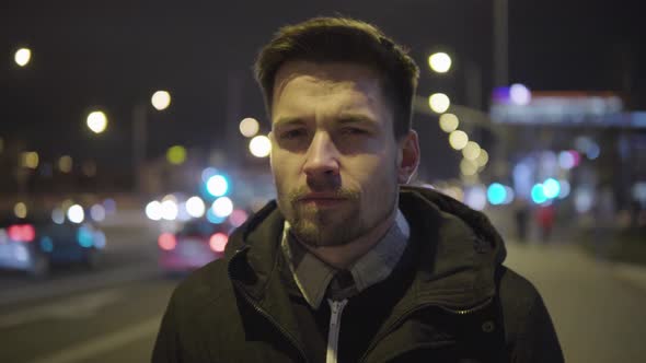 A Young Handsome Man Looks Seriously at the Camera in a Street in an Urban Area at Night - Closeup