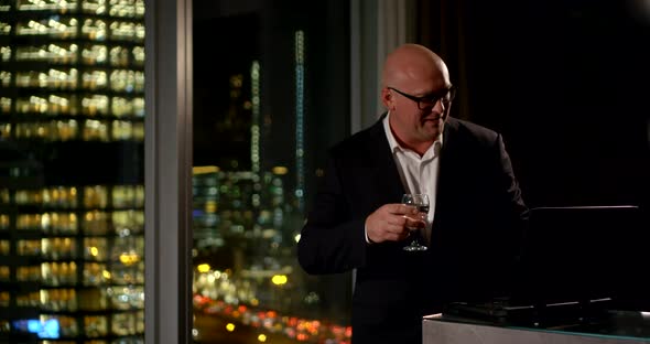 a Businessman in Glasses and Suit Cheerfully Communicates By Video Link on a Laptop Near a Large