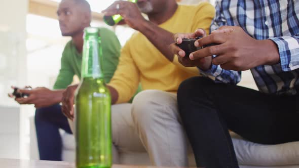 African american father and twin teenage sons sitting on couch playing game on tv and drinking beer