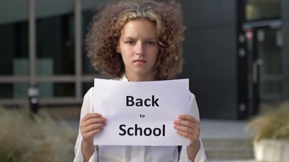 Cute Teenage Girl Stands with a Poster Back To School Near the School Building