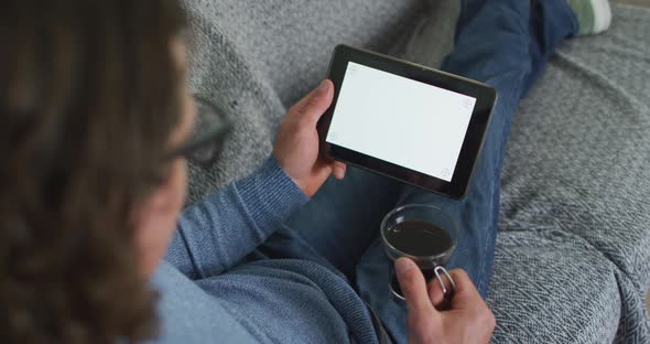 Caucasian man sitting on sofa in living room using tablet and drinking coffee