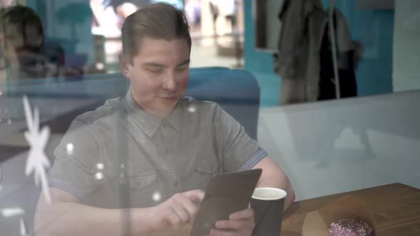 A Young Man is Sitting Behind a Glass in a Cafe with a Tablet in His Hands