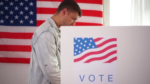 Young Smiling American Holds a Vote Sticker in Front of a Voting Booth. The Man Is Wearing a Denim