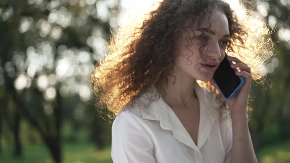 Busy Dissatisfied Young Caucasian Woman in Sunrays Talking on Phone Standing in Spring Summer Park