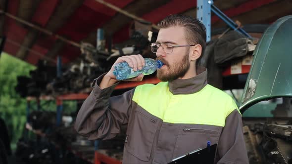 An Attractive Young Bearded Auto Mechanic in Glasses Stands at a Car Parts Warehouse and Drinks