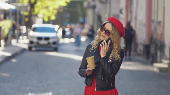 A Young Woman in the Middle of the Street Answers a Cell Phone Call and Drinks Takeaway Coffee