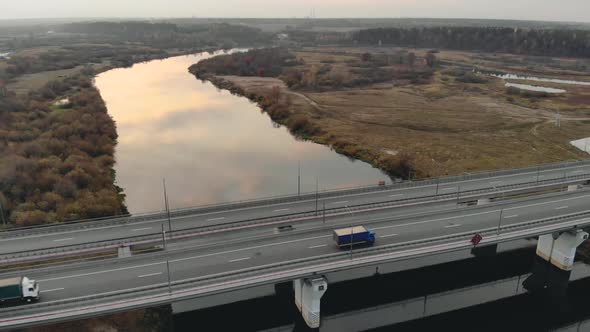 Aerial: Four-lane Road Bridge Over a River in Late Autumn with Two Trucks in Motion